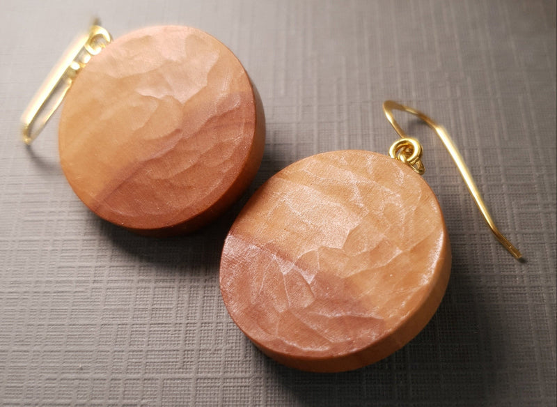 A pair of hammered round wooden drop earrings in a warm peachy-brown tone, suspended from gold fishhook wires, displayed on a textured gray surface.