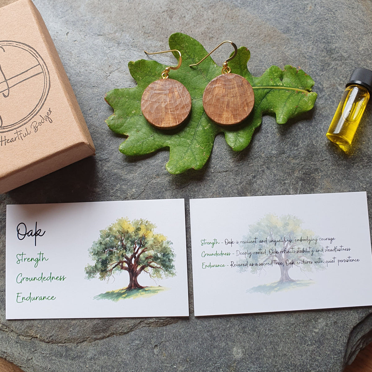 Round wooden ear hooks earrings displayed on green oak leaves, alongside a kraft paper box labeled 'The Healing Hands', a small bottle of essential oil, and two white cards with tree illustrations and text, all arranged on a round gray stone tray over a wood surface.