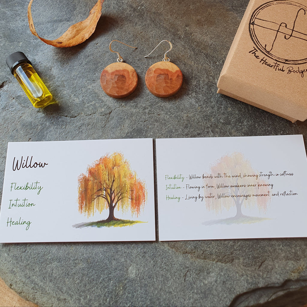 A styled flat-lay of a wellness product set on a round stone slab, featuring a pair of round wooden earrings, a small bottle of oil, a kraft paper box stamped with 'The Heartbeat Bridge', and two printed cards with autumn tree illustrations and text.