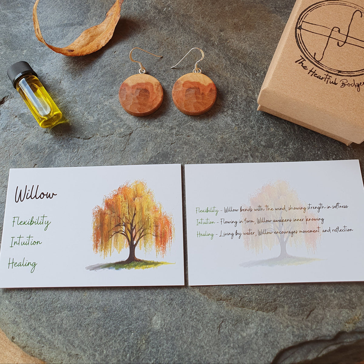 A styled flat-lay of a wellness product set on a round stone slab, featuring a pair of round wooden earrings, a small bottle of oil, a kraft paper box stamped with 'The Heartbeat Bridge', and two printed cards with autumn tree illustrations and text.