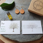 A styled flat-lay of a gift set on a round gray stone tray over a wooden surface. The set includes a small glass bottle of yellow oil, a green leaf, a pair of round wooden drop earrings, two printed cards with tree illustrations and text, and a kraft gift box with a logo.