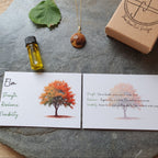 A jewelry and wellness gift set displayed on a round gray stone tray. The set includes a gold necklace with a brown wooden bead pendant, a small bottle of yellow oil, a green leaf, a kraft gift box stamped with 'The Heartful Bakes', and two printed cards featuring tree illustrations and text.
