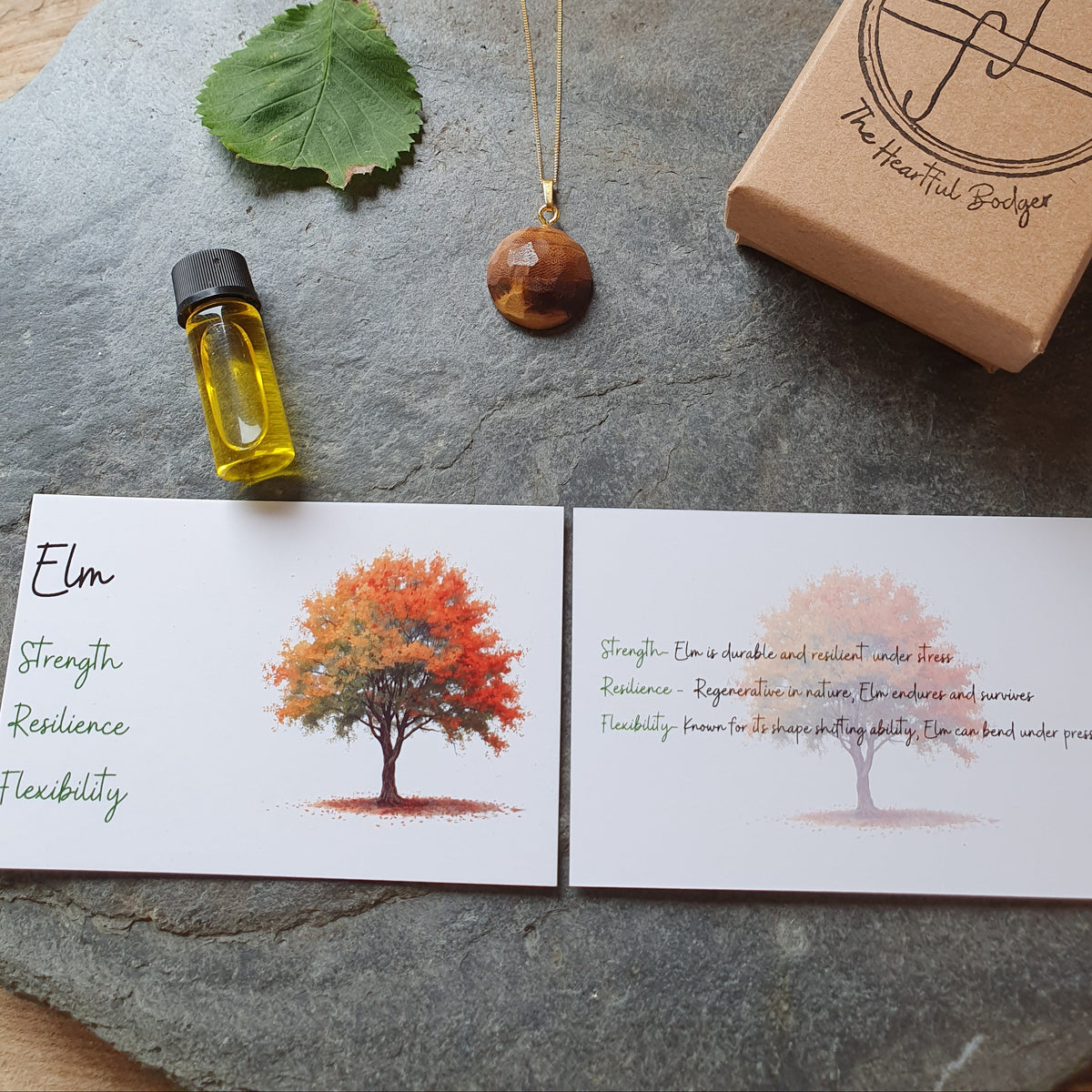 A jewelry and wellness gift set displayed on a round gray stone tray. The set includes a gold necklace with a brown wooden bead pendant, a small bottle of yellow oil, a green leaf, a kraft gift box stamped with 'The Heartful Bakes', and two printed cards featuring tree illustrations and text.