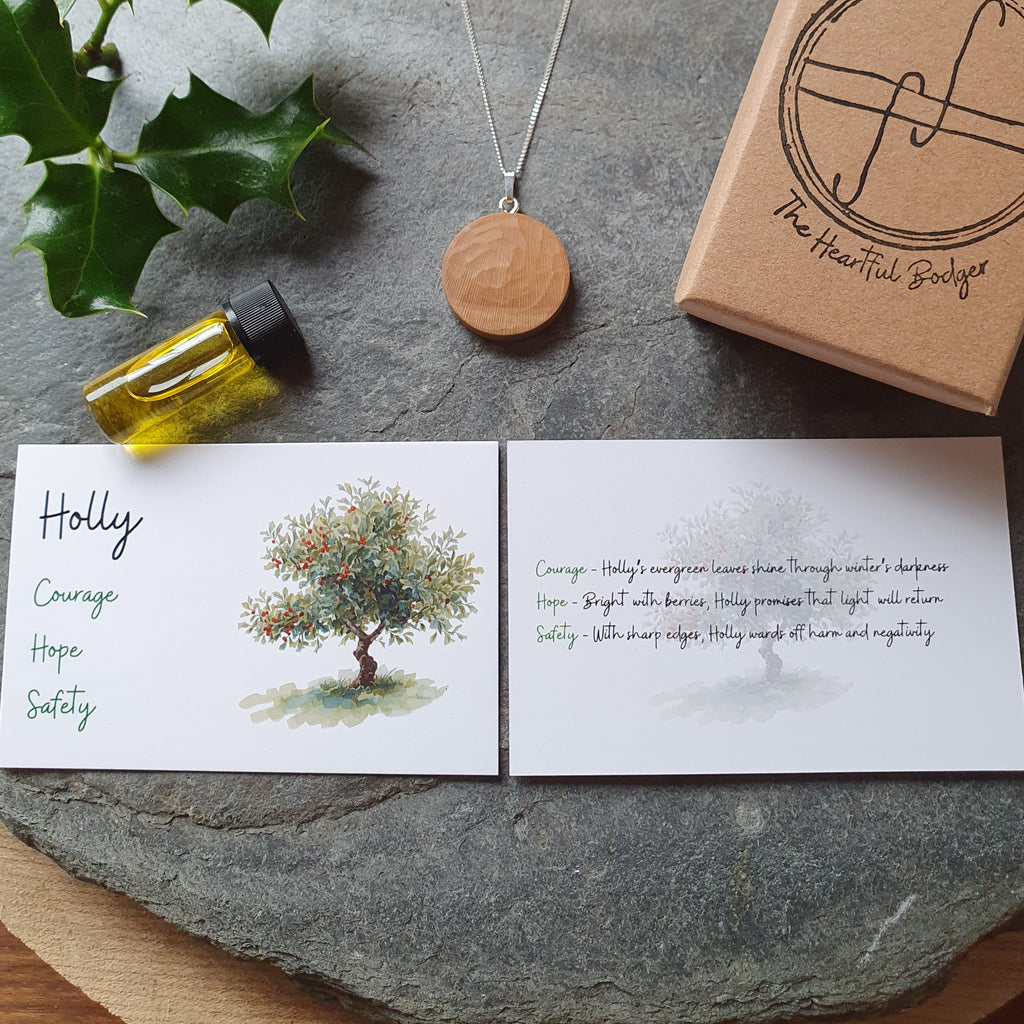 A styled flat-lay of a gift set on a round wooden board, featuring a silver necklace with a wooden pendant, a small kraft paper box with a logo, a small vial of oil, two printed cards with a tree illustration and text, and green holly leaves.